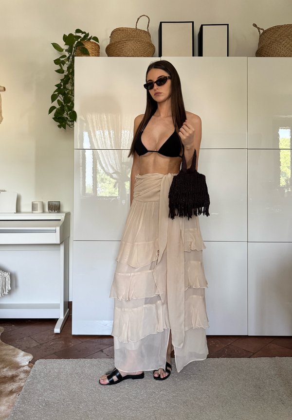Femeie purtând top de bikini negru, fustă lungă bej cu volane, sandale negre și ochelari de soare, ținând o geantă neagră țesută în interior.