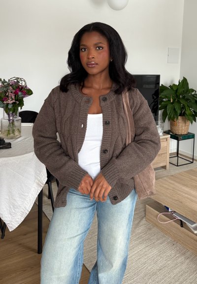 Mujer de cabello oscuro que lleva un cárdigan marrón, una camiseta sin mangas blanca y jeans azul claro, de pie en una sala de estar con plantas y una mesa.