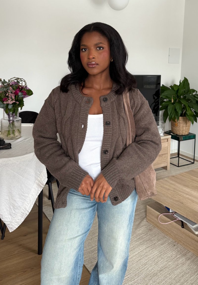 Donna con capelli scuri che indossa un cardigan marrone, una canotta bianca e jeans azzurri, in piedi in un soggiorno con piante e un tavolo.