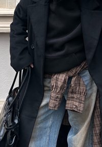 Schwarzer Wolltrenchcoat über einem schwarzen Hoodie, geschichtet mit einem braunen karierten Hemd, das um die Taille gebunden ist; Jeans mit unterschiedlichen Farbtönen. Schwarze Handtasche.