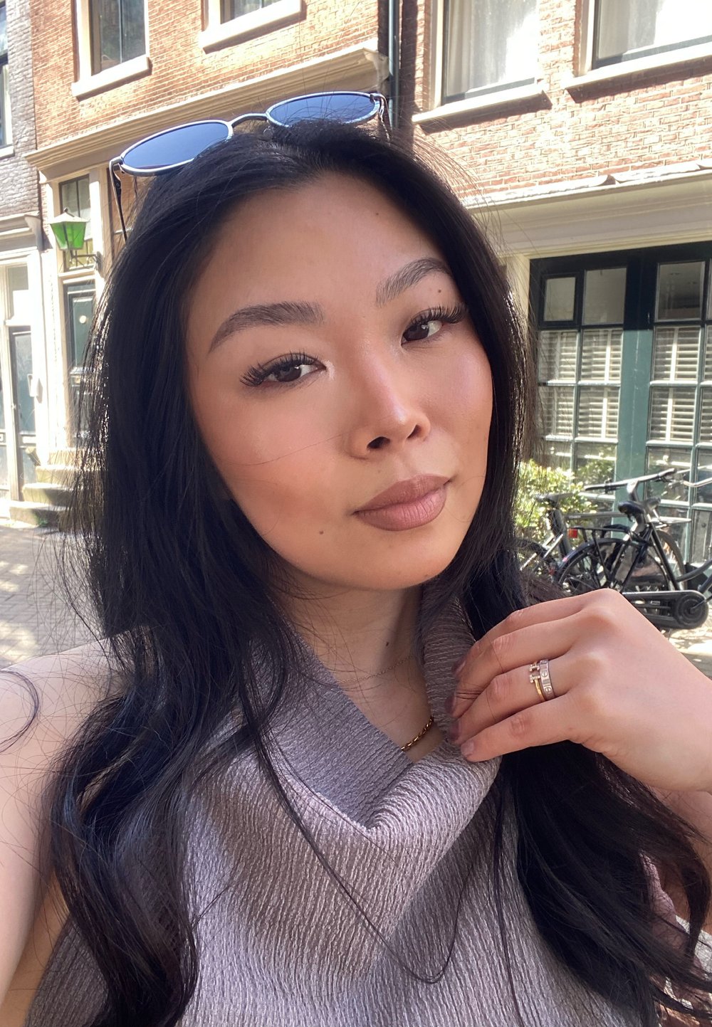 Primer plano de una persona con cabello largo y oscuro, vistiendo un top sin mangas de color gris claro con textura en relieve; se pueden ver un collar de cadena dorada y un anillo brillante.