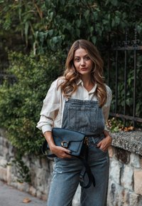 Des salopettes en denim affichent un délavé clair, des jambes larges et des poches, associées à un blouse crème à manches longues. Un sac à bandoulière noir avec des accents dorés est porté.