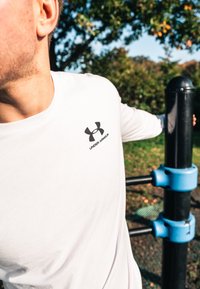 Balta ilga rankovė marškiniai su juodu Under Armour logotipu, prigludęs dizainas. Fone matoma žaliuojanti gamta ir lauko sporto įranga.