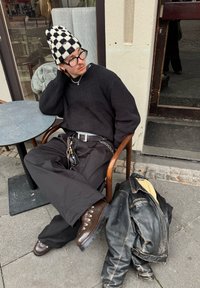 Črn pleteničast pulover, črne široke hlače, rjavi čevlji z vezalkami, karirasta kapa, srebrni dodatki in obrabljen črn usnjen jakno, ki leži v bližini.
