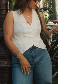 Hvid linned ærmeløs top med V-udskæring og knapper, parret med højtaljede blå denim jeans. Accessories består af armbånd og en håndtaske.
