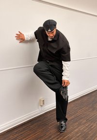 Chemise noire à manches courtes portée sur un t-shirt blanc à manches longues, pantalon noir et chaussures noires vernies. La personne plie la jambe et s'appuie contre le mur.