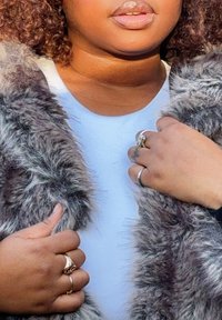 Gilet en fausse fourrure grise texturée porté sur un haut bleu clair. Les mains sont ornées de plusieurs bagues, présentant divers motifs et textures.