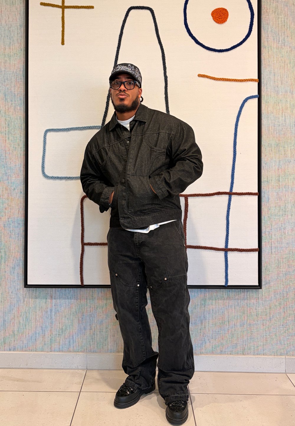 Veste et pantalon en denim noir à coupe décontractée, superposés sur une chemise blanche, accompagnés de baskets noires. Debout devant une œuvre murale abstraite.