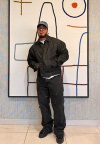 Veste et pantalon en denim noir à coupe décontractée, superposés sur une chemise blanche, accompagnés de baskets noires. Debout devant une œuvre murale abstraite.