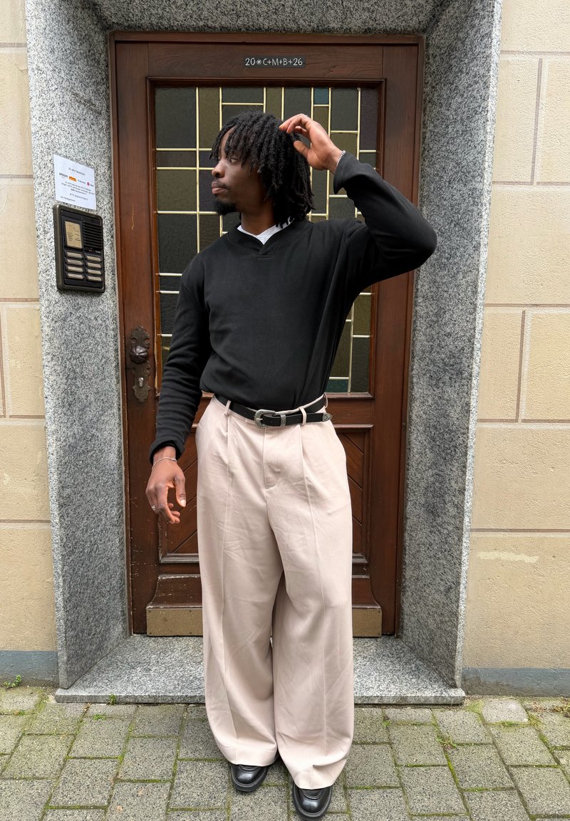 Homme avec dreadlocks portant un pull noir et un pantalon beige large, debout devant une porte en bois encadrée par des murs en pierre et en brique.