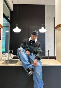 Ung mann som har på seg en svart og grønn genser og revne jeans sitter på en kjøkkenbenk og justerer buksebeinet under to hengende lys.