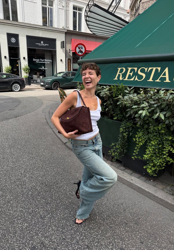 Una mujer sonriente posa con un bolso marrón junto a una jardinera frondosa y un toldo verde de restaurante en una calle de la ciudad.