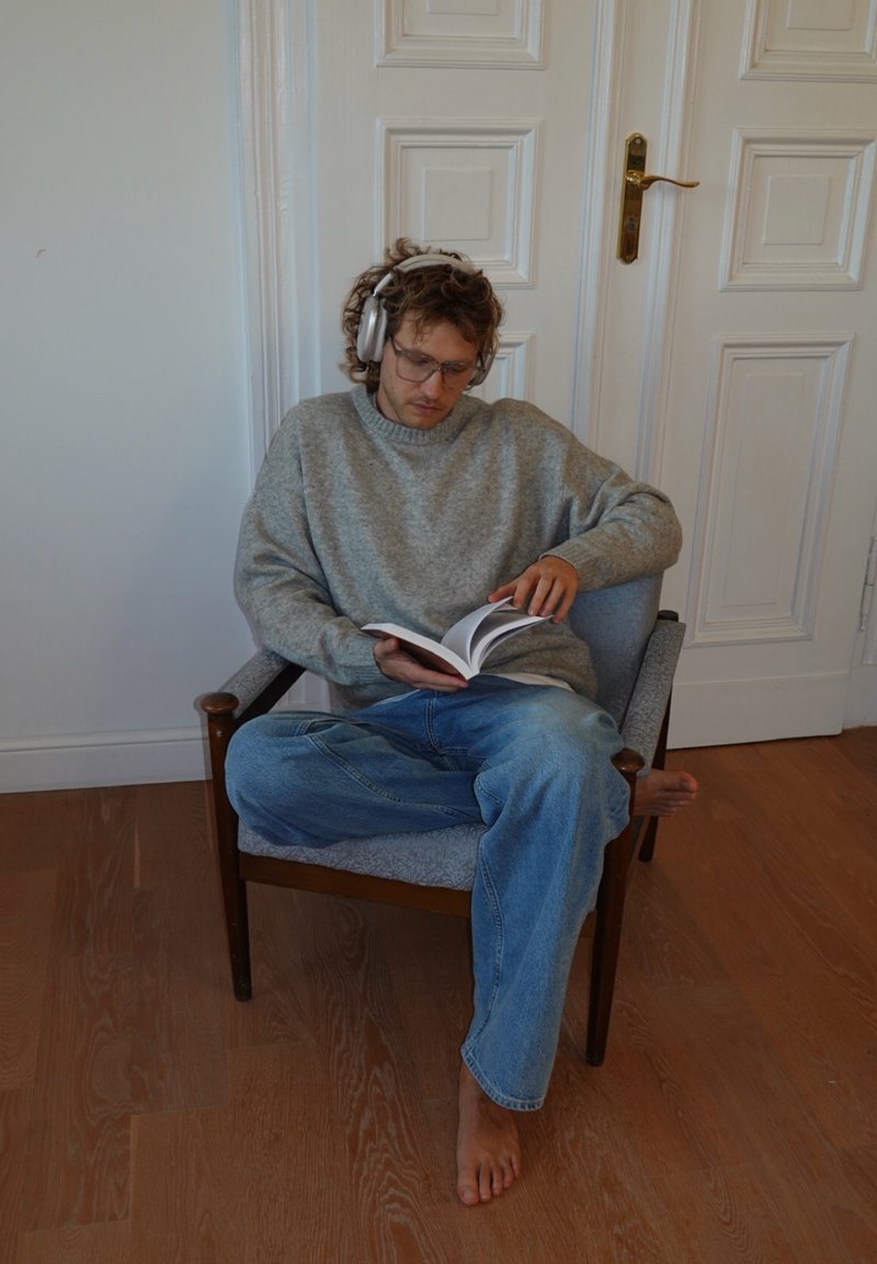 Personne assise dans un fauteuil rembourré bleu clair, portant un pull gris et un jean en denim, lisant un livre avec un casque audio.
