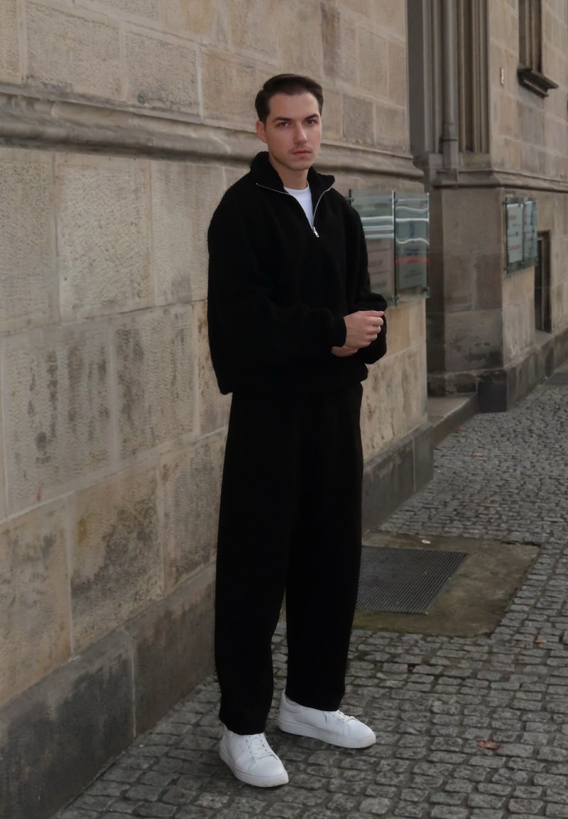 Pull noir à fermeture éclair, texture douce, associé à un pantalon noir ample. Des baskets blanches complètent la tenue. Mur en pierre neutre en arrière-plan.