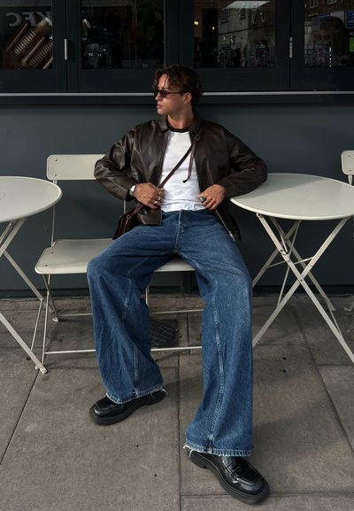 Hombre con gafas de sol, chaqueta de cuero, camisa blanca y vaqueros anchos sentado en una silla de café al aire libre entre mesas blancas, mirando hacia un lado.