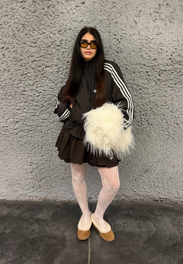 Une femme portant des lunettes de soleil, une veste noire à rayures, une jupe, des collants en dentelle et des chaussures bordées de fourrure pose devant un mur gris, un sac blanc duveteux à la main.