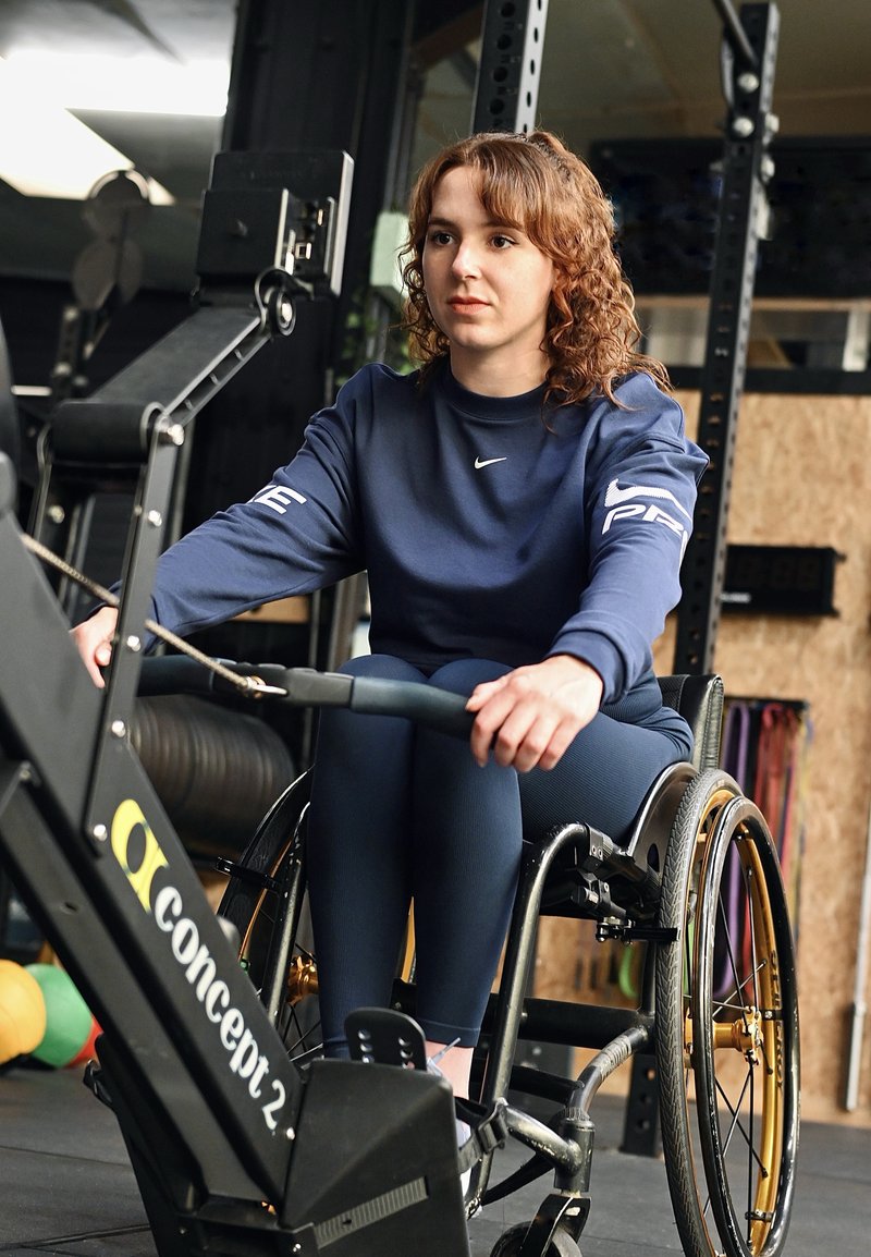 Donna in sedia a rotelle che utilizza una macchina per il vogatore, concentrata sull'allenamento in una palestra con attrezzature fitness sullo sfondo.