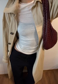 Beżowy trencz z czarnymi guzikami na dopasowanej białej koszuli, zestawiony z czarnymi spodniami i plecioną bordową torebką.