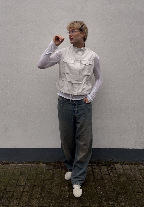 Personne portant un gilet léger, un haut transparent à manches longues, un jean ample et des baskets blanches, debout contre un mur uni et regardant sur le côté.
