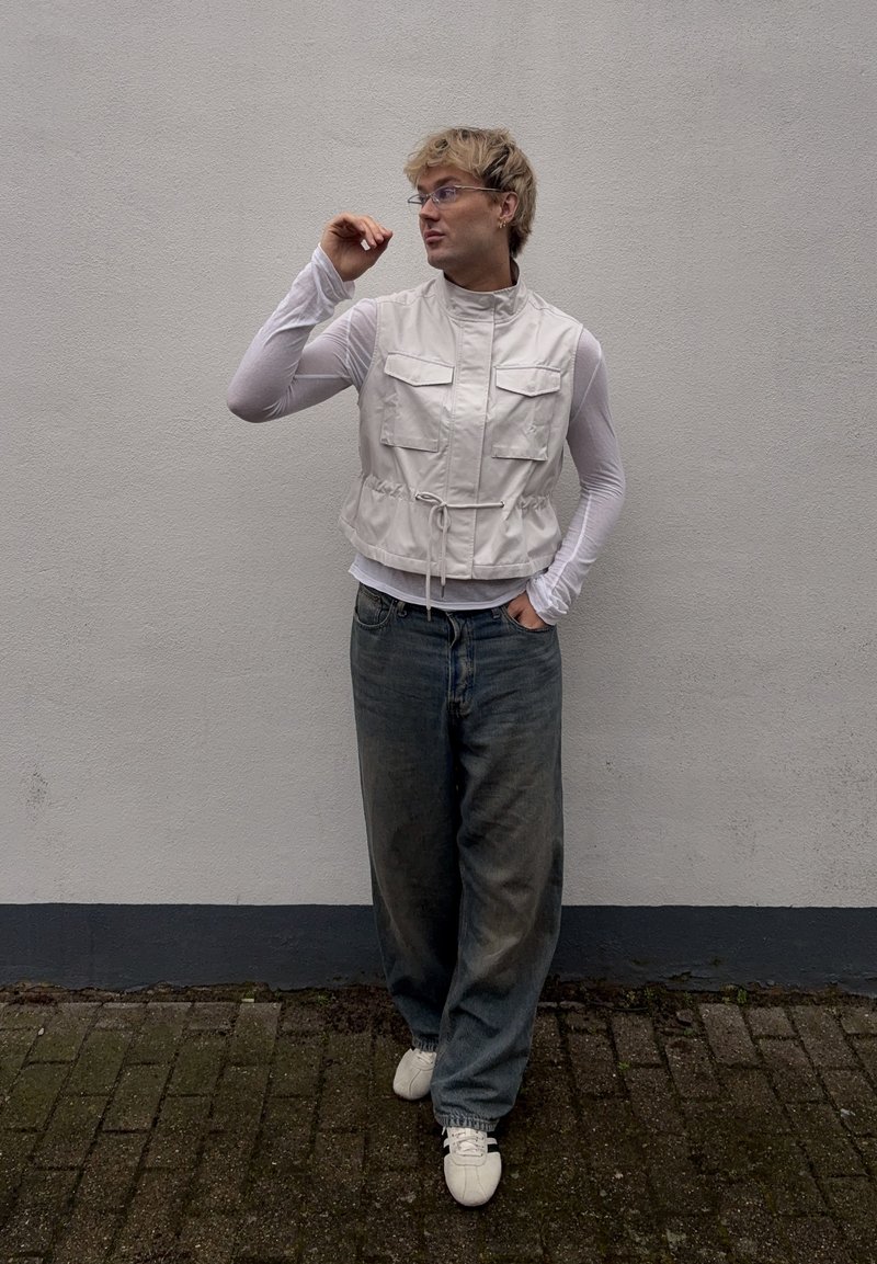 Personne portant un gilet léger, un haut transparent à manches longues, un jean ample et des baskets blanches, debout contre un mur uni, regardant sur le côté.