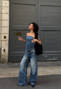 Kvinna med lockigt hår iklädd en denim tubtopp och vida jeans står på gatan nära en grå dörr, och håller en svart pälsig väska på axeln.