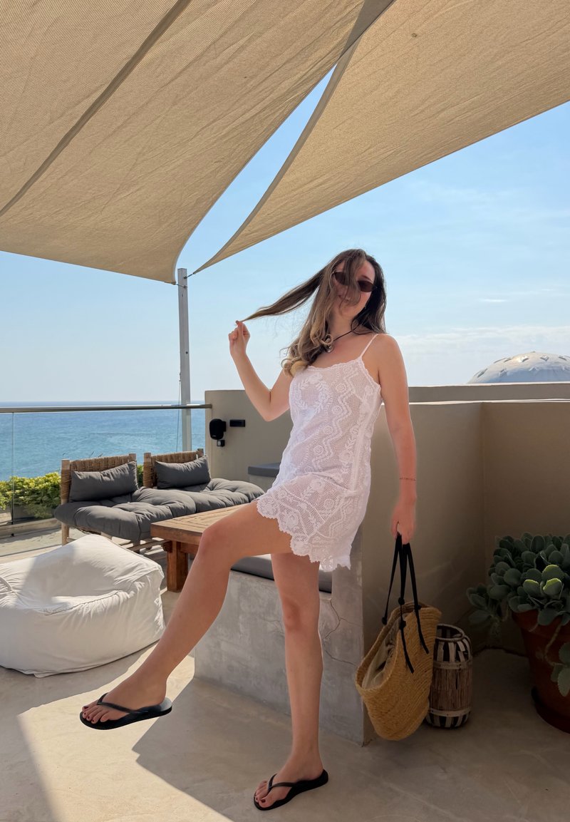 Γυναίκα με λευκό δαντελένιο φόρεμα και γυαλιά ηλίου κρατάει τα μαλλιά και μια ψάθινη τσάντα, στέκεται σε ηλιόλουστη βεράντα με θέα στον ωκεανό και σκιασμένη περιοχή καθιστικού.