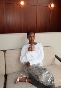 Vit kabelstickad tröja bärs med en glittrande silverkjol. Sitter på en beige soffa och håller i en genomskinlig drink med ett sugrör. Träpanel bakom.