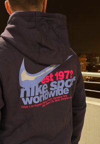 Svart hettegenser med en stor blå Nike-logo, rosa "est 1972," og teksten "nike sport worldwide" i grått, med bynavn listet under.