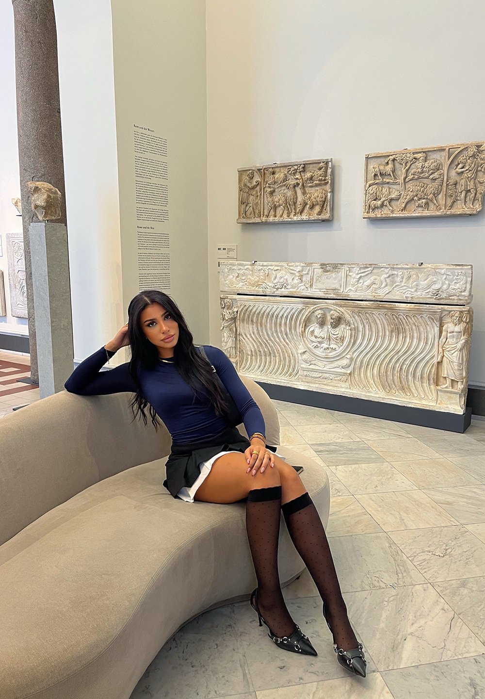 Kurvad beige soffa, en person i en marinblå långärmad topp och svart kjol sitter bredvid en vit stenskulptur, med polerade mörka skor och mönstrade strumpor.