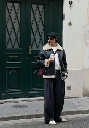 Chaqueta de cuero negra con cuello de borreguito, suéter blanco, pantalones oscuros de pierna ancha, boina negra y gafas de sol; sosteniendo una taza de café.