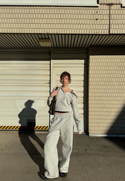 Sudadera corta con cremallera de color gris claro, con puños acanalados y pantalones de chándal sueltos a juego. La modelo sostiene un pequeño bolso marrón, de pie contra una pared texturizada.
