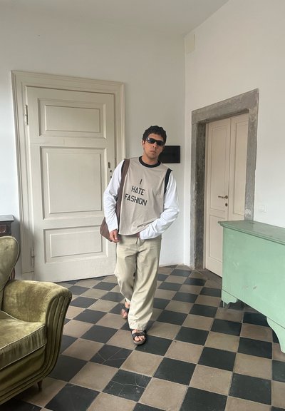 Top sin mangas de color gris claro con el texto "ODIO LA MODA" sobre una camiseta de manga larga blanca, pantalones beige y sandalias negras. Habitación con suelo de damero.