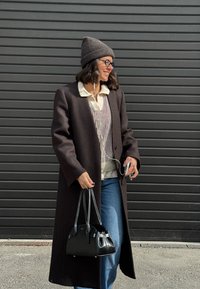 Kobieta w ciemnym długim płaszczu, czapce beanie i okularach, trzymająca czarną torebkę i telefon z słuchawkami, stojąca przy szarej metalowej ścianie.