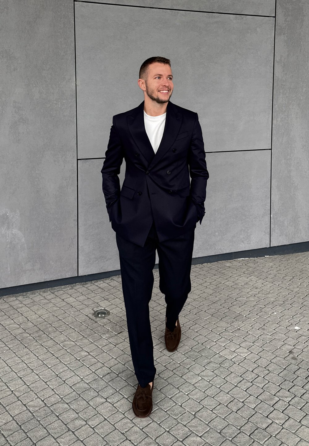 Férfi fekete öltönyben, fehér ingben és barna cipőben sétál a csempézett járdán, mosolyogva, oldalra nézve egy szürke fal előtt.