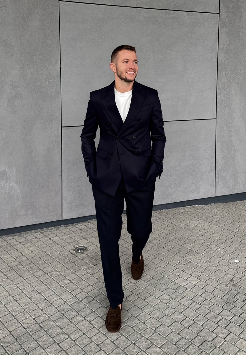 Man walking on tiled pavement wearing a black suit, white shirt, and brown shoes, smiling and looking to the side against a grey wall.