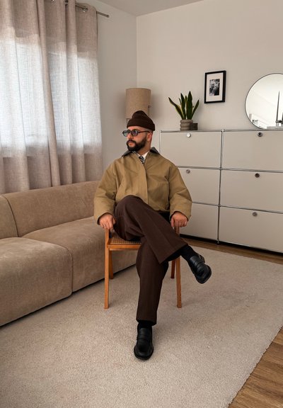 Hombre con gafas y gorro, vestido con chaqueta beige y pantalones marrones, sentado con las piernas cruzadas en una silla de madera en una sala de estar moderna.