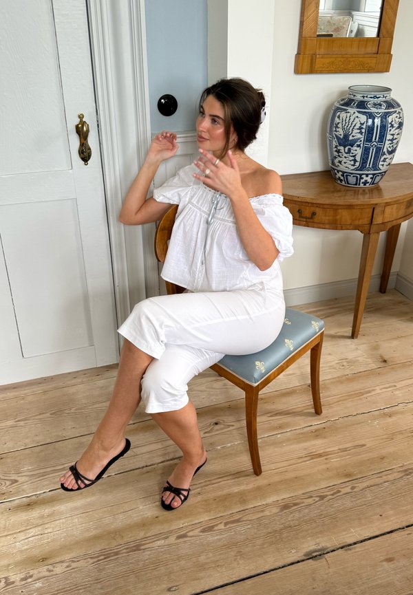 Femme en haut blanc à épaules dénudées et pantalon blanc assise sur une chaise, touchant ses cheveux, avec un vase en céramique sur une table en arrière-plan.