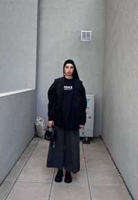 Μαύρη oversized φούτερ με το λογότυπο "NIKE", ταιριαστό μαύρο γιλέκο, γκρι παντελόνι με φαρδιά πόδια, μαύρες γαλότσες, μικρή τσάντα και μαύρο μαντήλι.