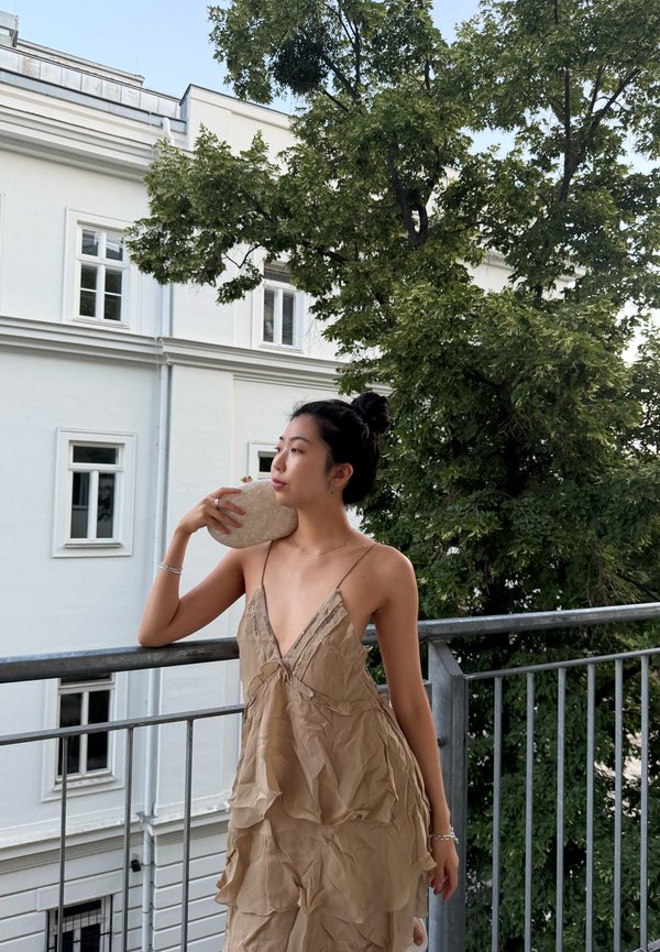 Junge Frau in einem beigen, geschichteten Kleid steht auf einem Balkon und hält eine helle Stein-Clutch nahe an ihrem Gesicht, im Hintergrund ein Baum und ein weißes Gebäude.