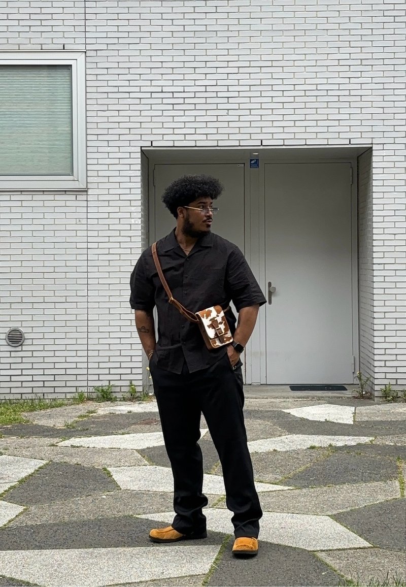 Μαύρο κοντομάνικο πουκάμισο και μαύρο παντελόνι φορεμένα με καφέ παπούτσια. Καφέ τσάντα χιαστί με λευκό και καφέ σχέδιο. Στεκόμενος σε πλακοστρωμένο έδαφος.