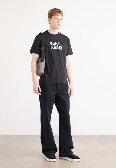 Camiseta de algodón negra con logo blanco, combinada con pantalones vaqueros de pierna ancha azul oscuro y zapatillas negras. Bolso cruzado gris con acentos negros.