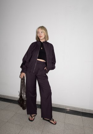 Mujer que lleva una chaqueta morado oscuro y pantalones de pierna ancha, top corto negro, sosteniendo una bolsa con ojales de metal, de pie contra una pared blanca.