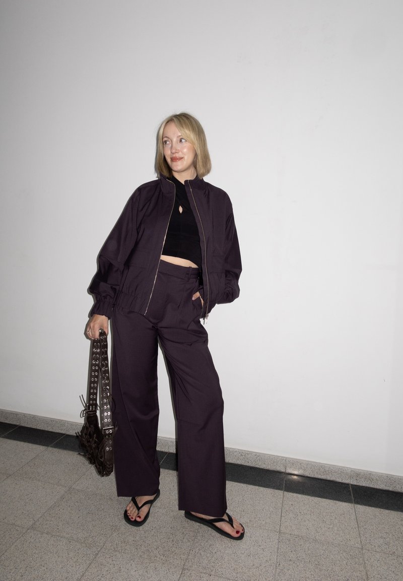 Femme portant une veste violet foncé et un pantalon à jambes larges, un crop top noir, tenant un sac avec des œillets métalliques, debout contre un mur blanc.