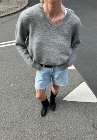 Sivi pleteni pulover s V-izrezom, svetloplave jeans kratke hlače s raztrganimi robovi in črne čevlje s konicami. Stoji na sivi asfaltni površini.