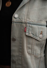 Světle modrá denimová bunda se stříbrnými knoflíky, přední kapsou a malou červenou etiketou Levi's na bočním švu. Hladká textura a klasický design.