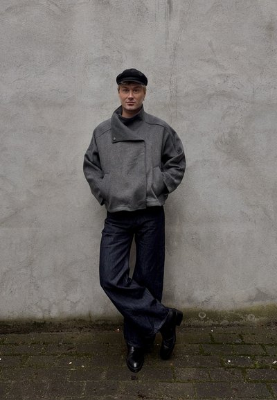 Persona joven con chaqueta gris, gorra negra, pantalones anchos oscuros y botas negras, de pie contra una pared gris texturizada con las manos en los bolsillos.