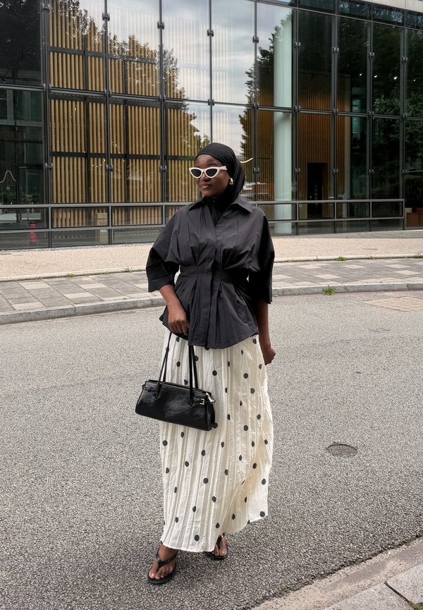 Donna con top nero, gonna bianca a pois, occhiali da sole bianchi, hijab nero e sandali, che tiene una borsa nera in una strada di città.