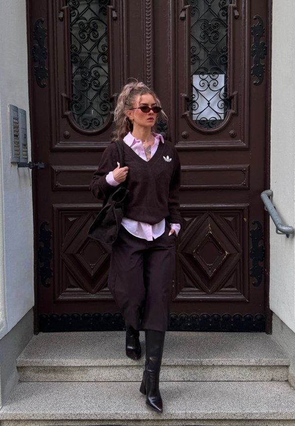 Femme portant des lunettes de soleil, un pull noir, une chemise rose, un pantalon noir et des bottes descend des marches en pierre devant des portes doubles ornées foncées.