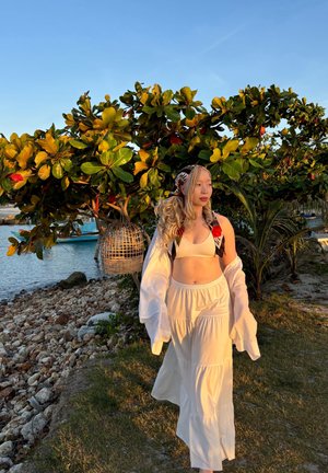 Falda maxi blanca y con volantes, y top corto con mangas largas y fluidas. El fondo incluye un exuberante follaje verde y una linterna colgante tejida.