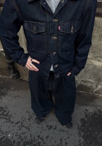 Jeans jakna z dvema žepoma na prsih in vidnimi šivi, kombinirana s širokimi jeans hlačami. Obe sta temno modri barve in imata sproščen kroj.